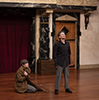 Lear, wearing black shirt and gray pants, stands stalwart with head raised while the Fool in houndstooth cap and wrapping Lear's brown overcoat around herself kneels on the otherwise bare stage. 